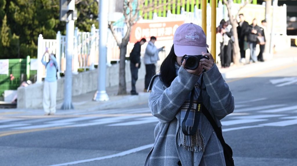 A girl wearing a pink hat and a gray plaid coat is on a street, taking pictures of herself with a black camera.