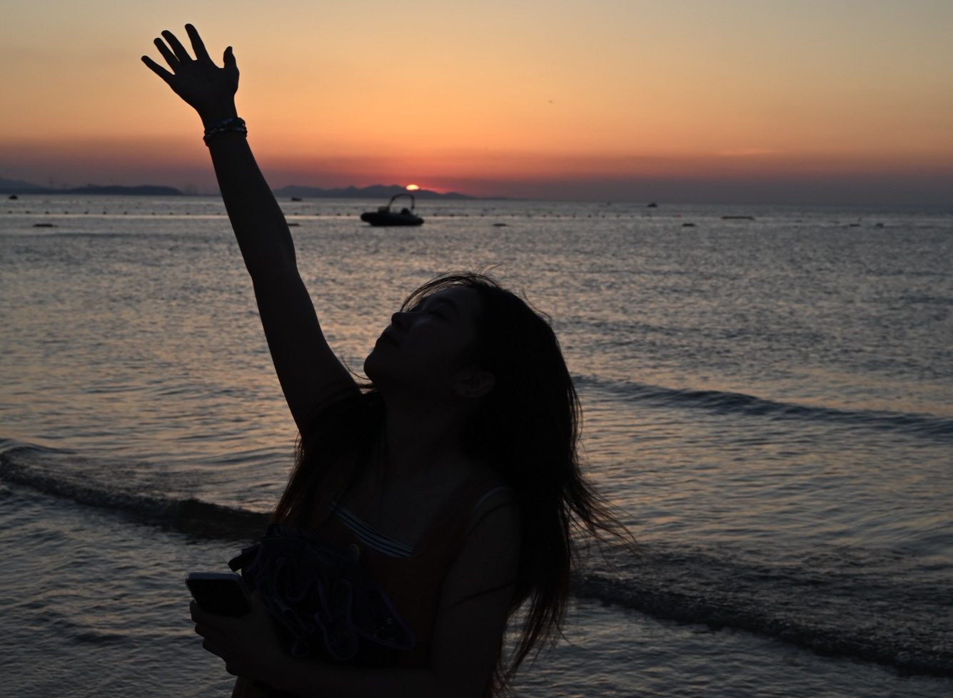 A girl, seen in silhouette against the light, raises her right hand to feel the wind blowing through her fingertips; the background features the deep blue sea and a sunset sky blending yellow and blue hues.